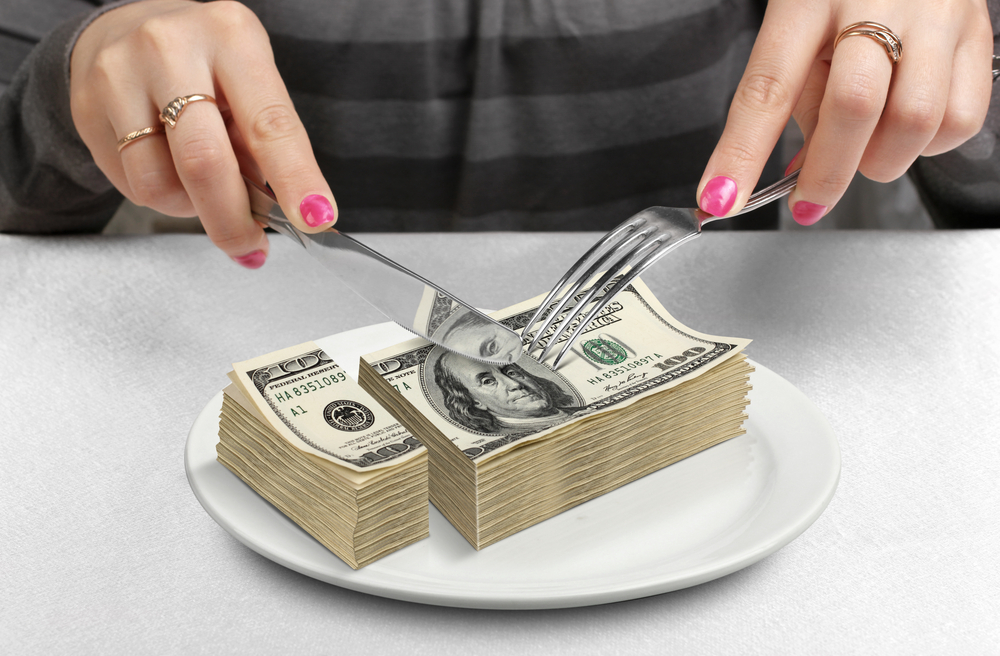Close-up of woman's hands as she cuts up a stack of $100 bills with a knife and fork.