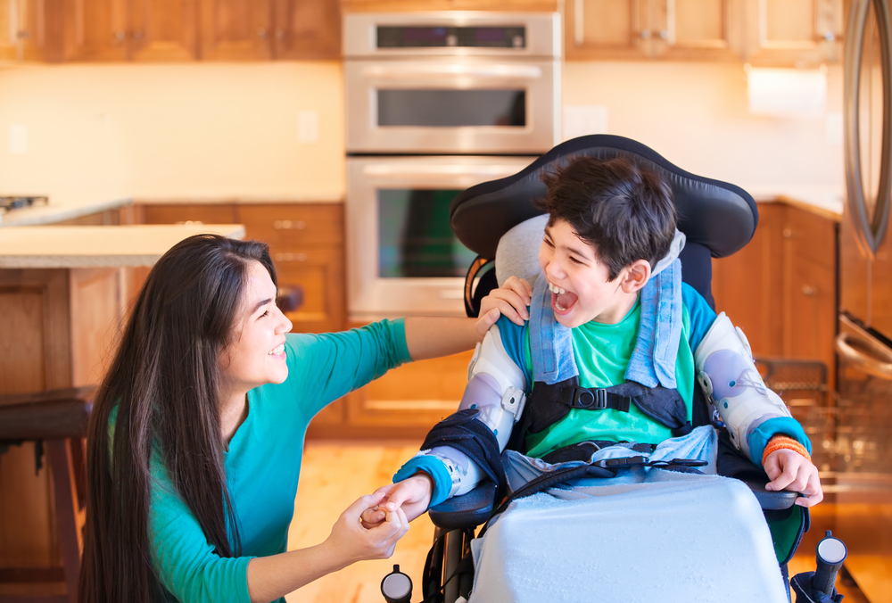 Boy with disability using wheelchair laughing with family caregiver in kitchen.