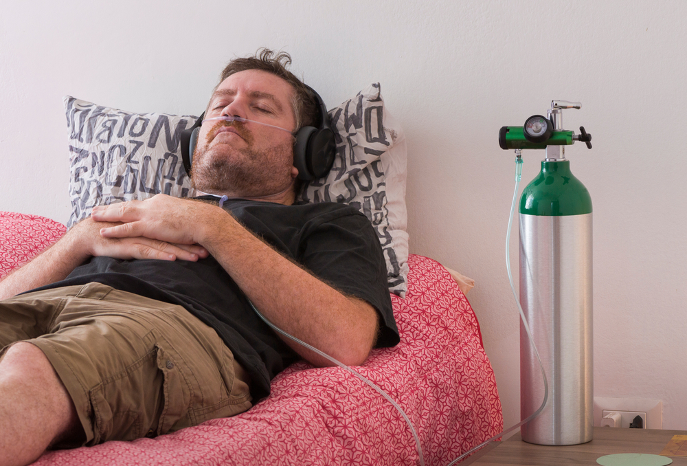 A man lies on his bed resting and breathing with the help of a portable oxygen tank.