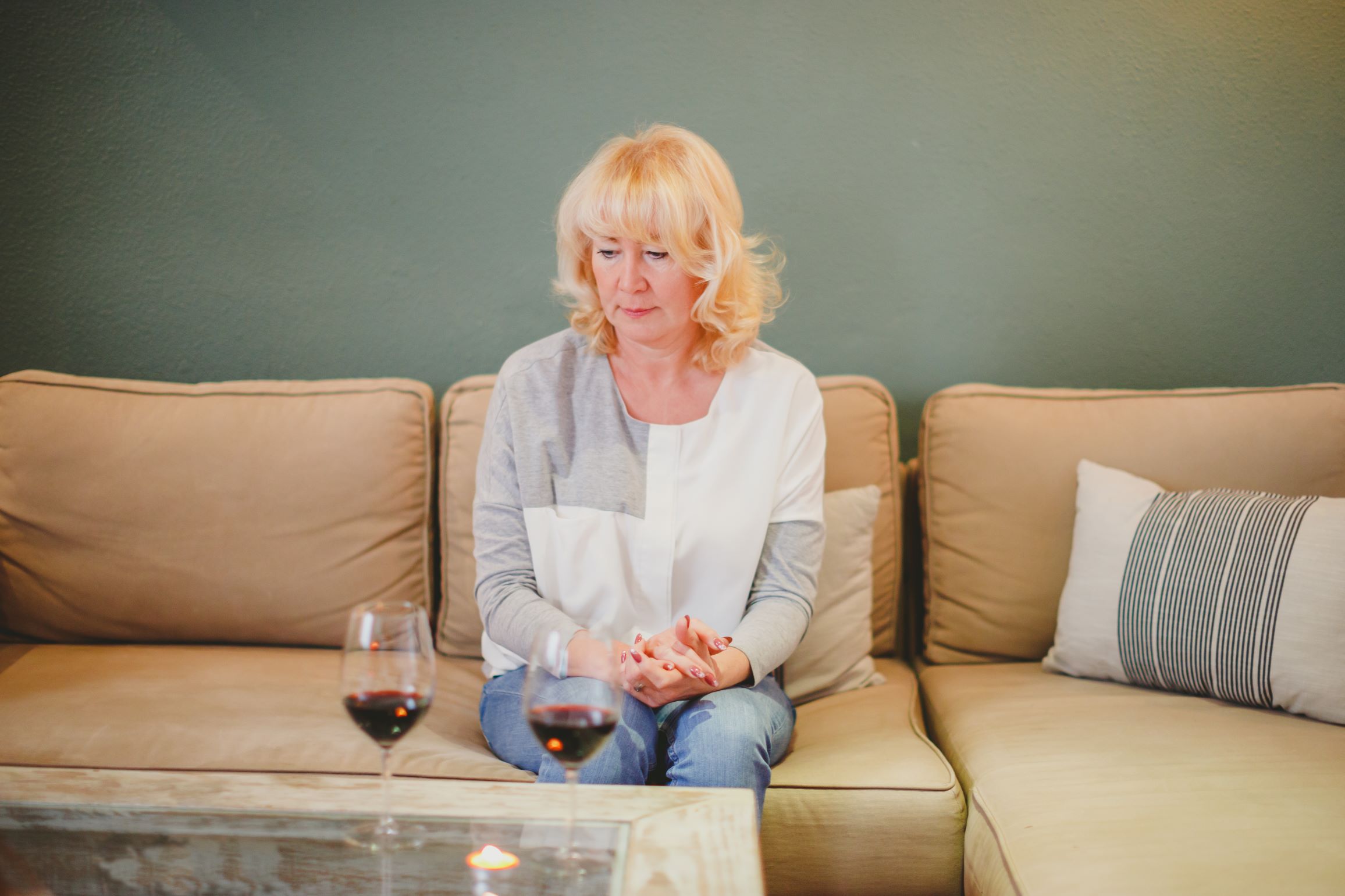 Middle-aged woman sits looking disappointed and waiting alone on couch with two glasses of wine on the table in front of her.
