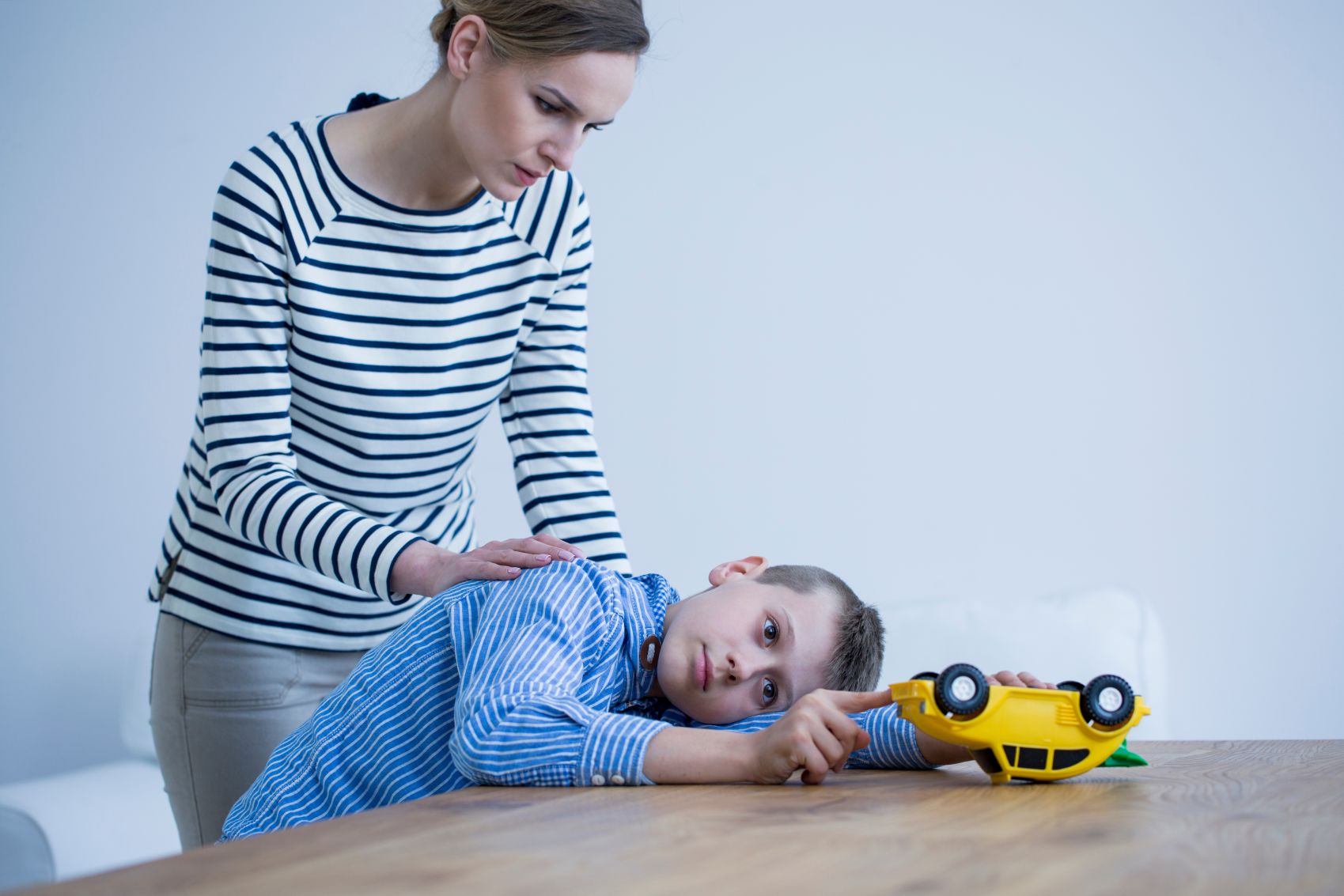 Concerned mother stands over son who is looking listlessly at toy car.